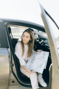 Young woman sitting in car holding a map, planning her journey with the door open.